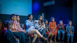 A group of children and young women participating in a panel discussion indoors.