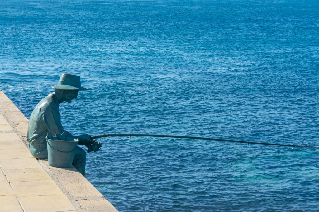 A bronze statue of a fisherman sitting by the ocean, casting a fishing line into the blue sea.