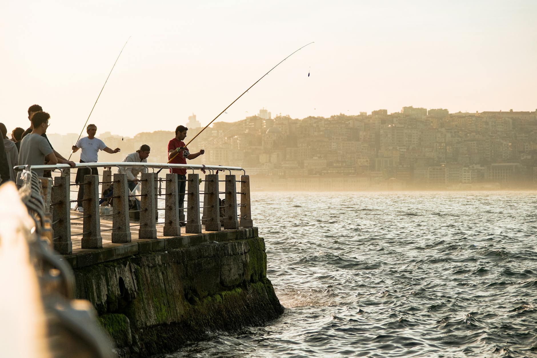 A group of men fishing on a pier during sunset, city skyline in background.