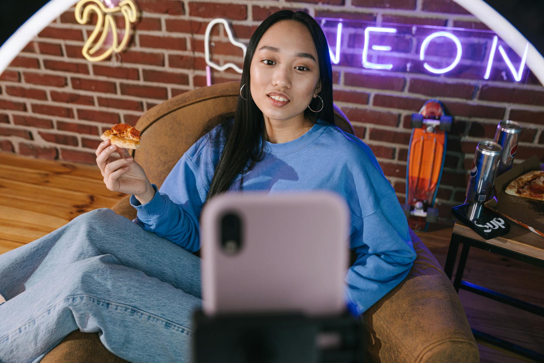 Young Asian woman streaming video while enjoying pizza in a cozy setup with neon lights.
