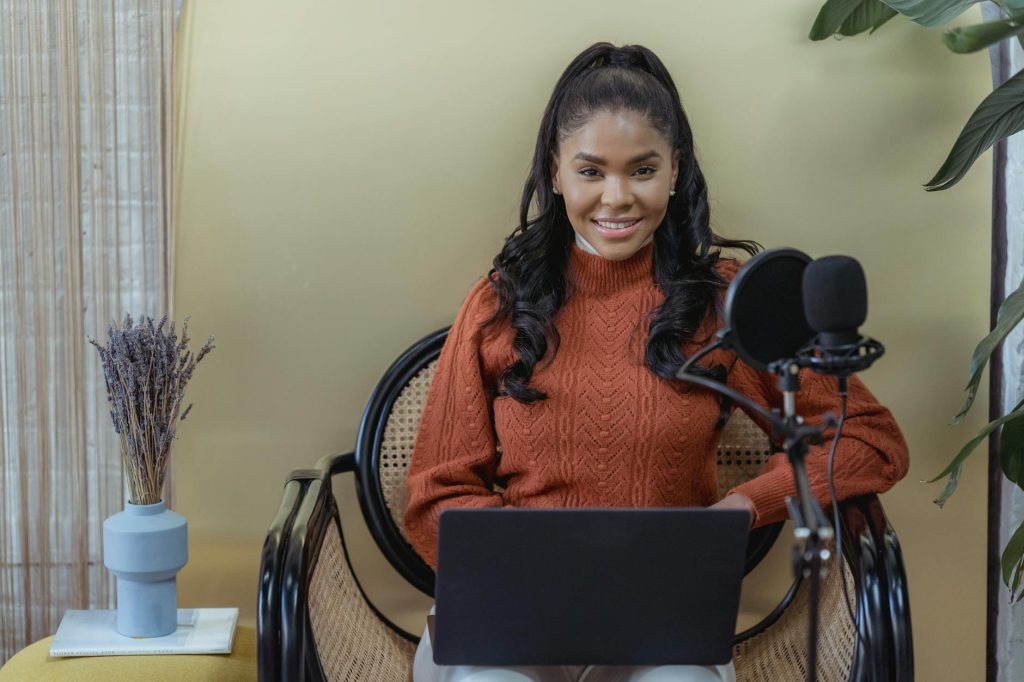 Happy young African American female blogger smiling at looking at camera while using laptop in studio during podcast recording