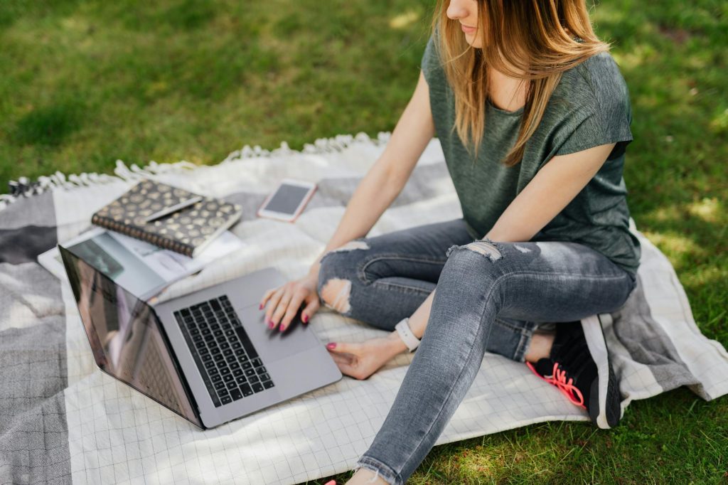 From above of crop young female student in casual clothes sitting with laptop on blanket placed on green park lawn and doing homework on sunny day