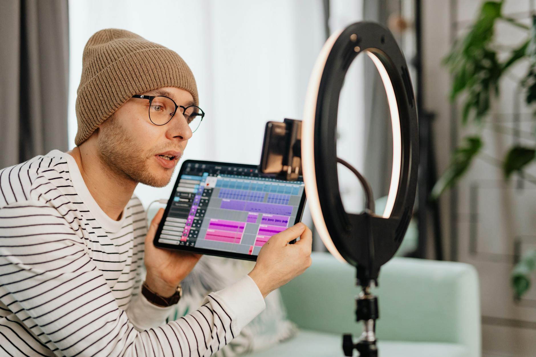 A young man wearing glasses and a beanie records a video indoors using a tablet and ring light.