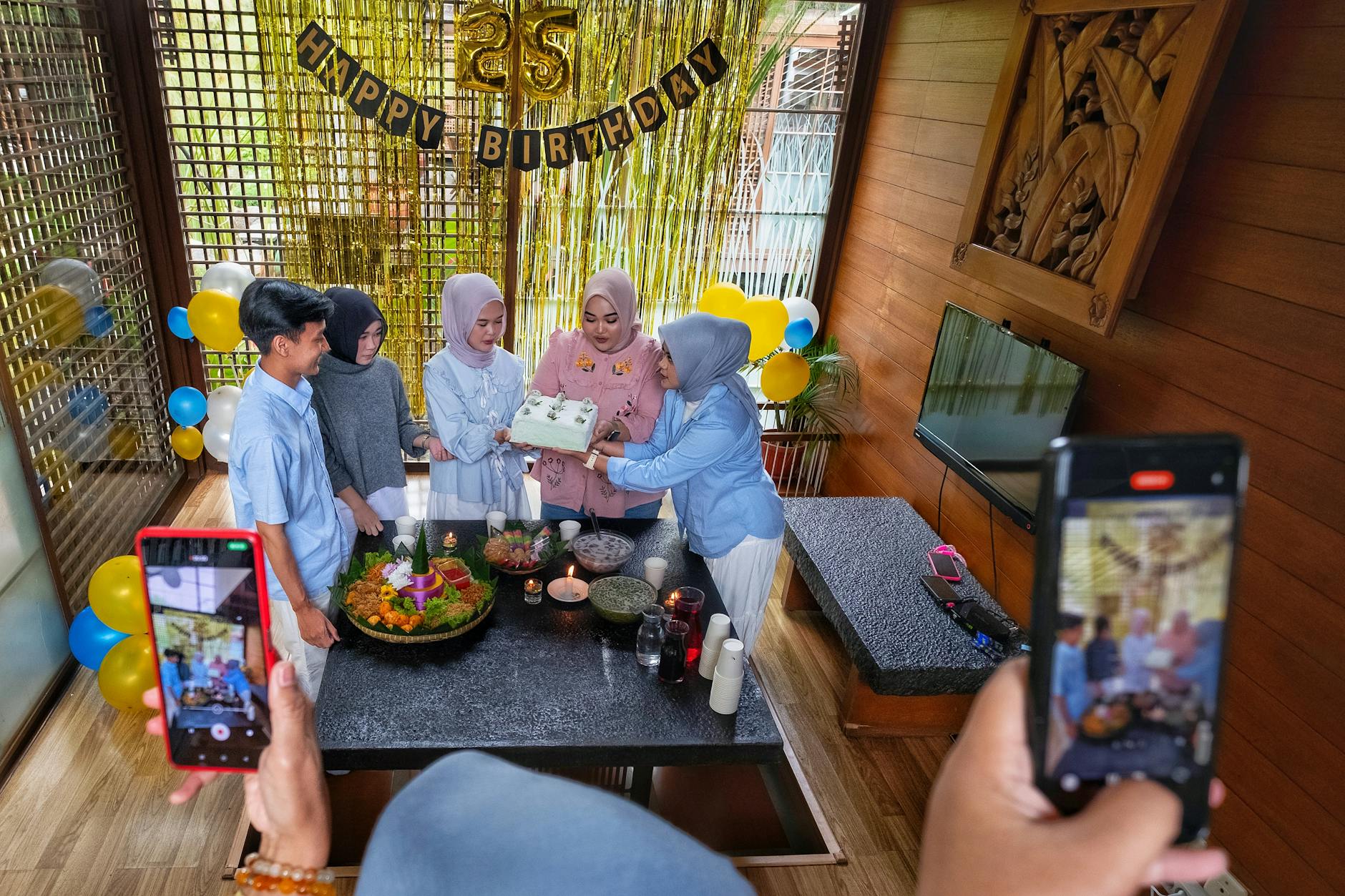 Group celebrating 25th birthday indoors with cake and balloons.
