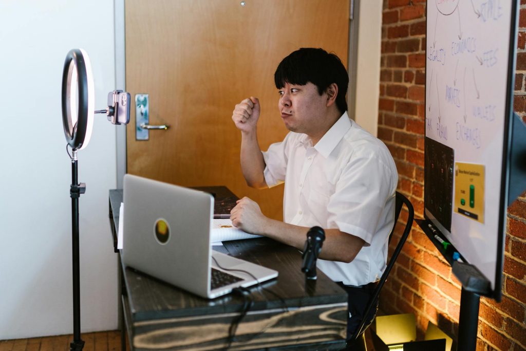 Asian man in white shirt conducts an online meeting with a laptop and ring light.