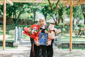 A young Muslim woman celebrating graduation in Shah Alam, Malaysia, outdoors in a park with flowers.