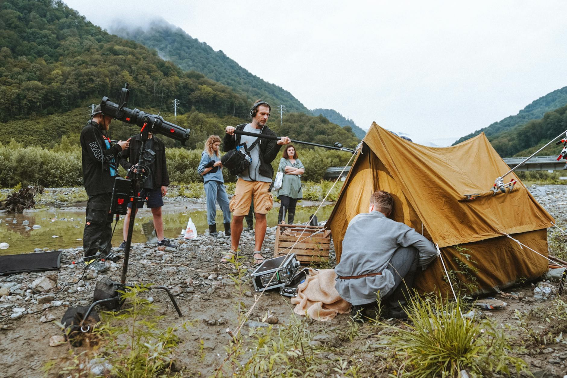 A film crew working with equipment and a tent by a river in a mountainous location.