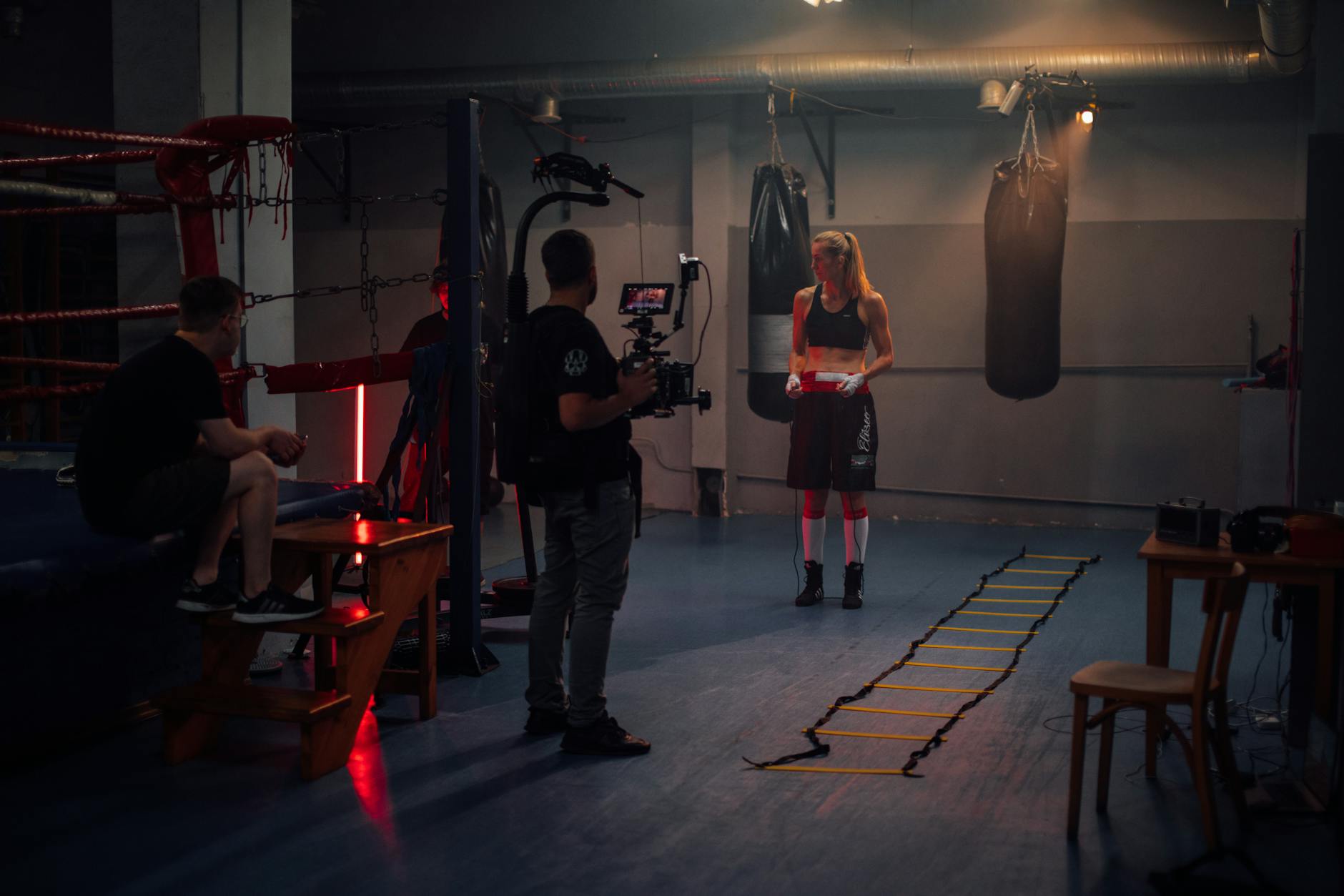 Female boxer preparing in a dimly lit boxing gym with a film crew.