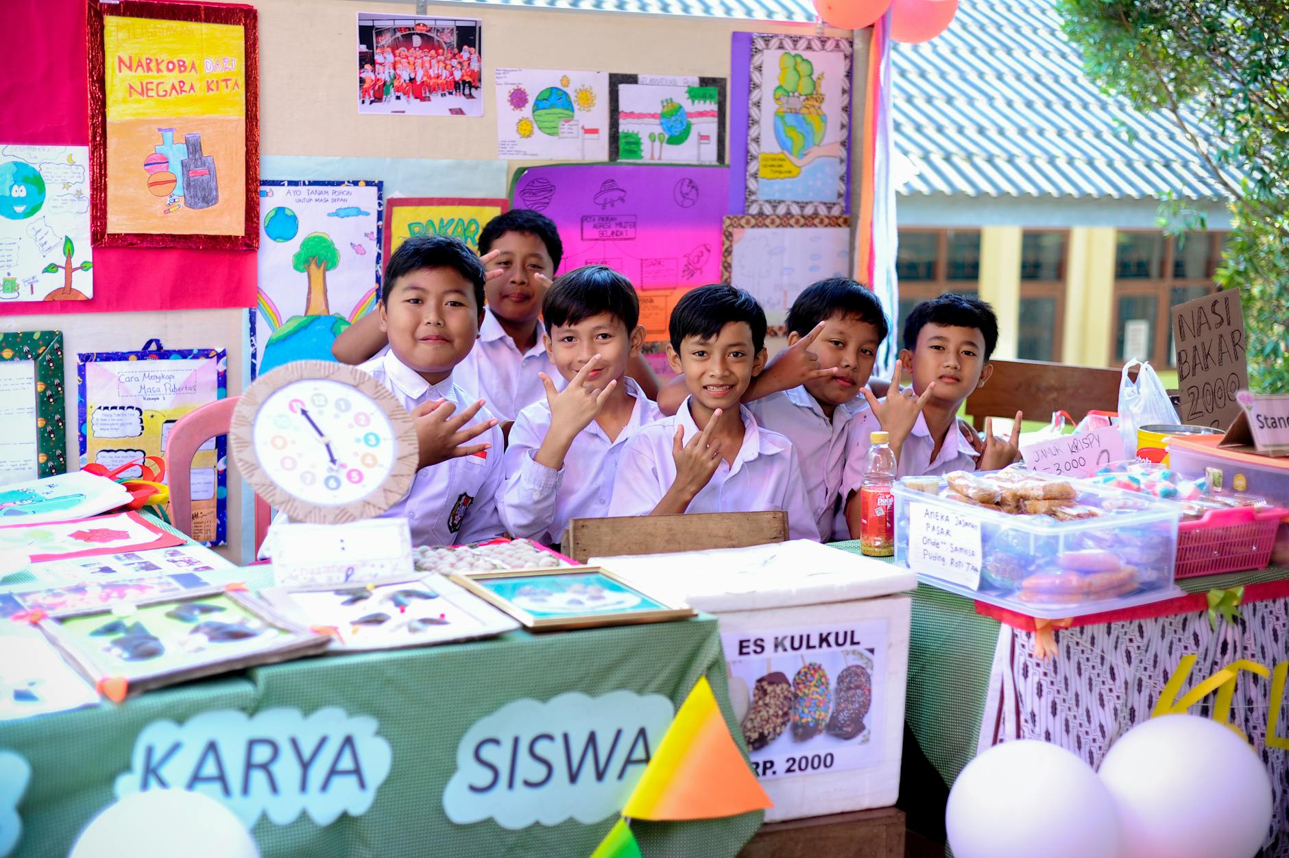 Students pose at a vibrant school exhibition, showcasing projects and crafts with smiles and gestures.