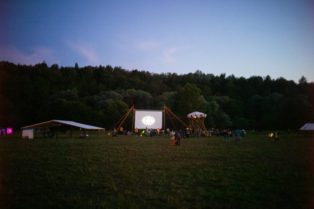 A twilight outdoor movie event in a meadow with people gathered around a screen.