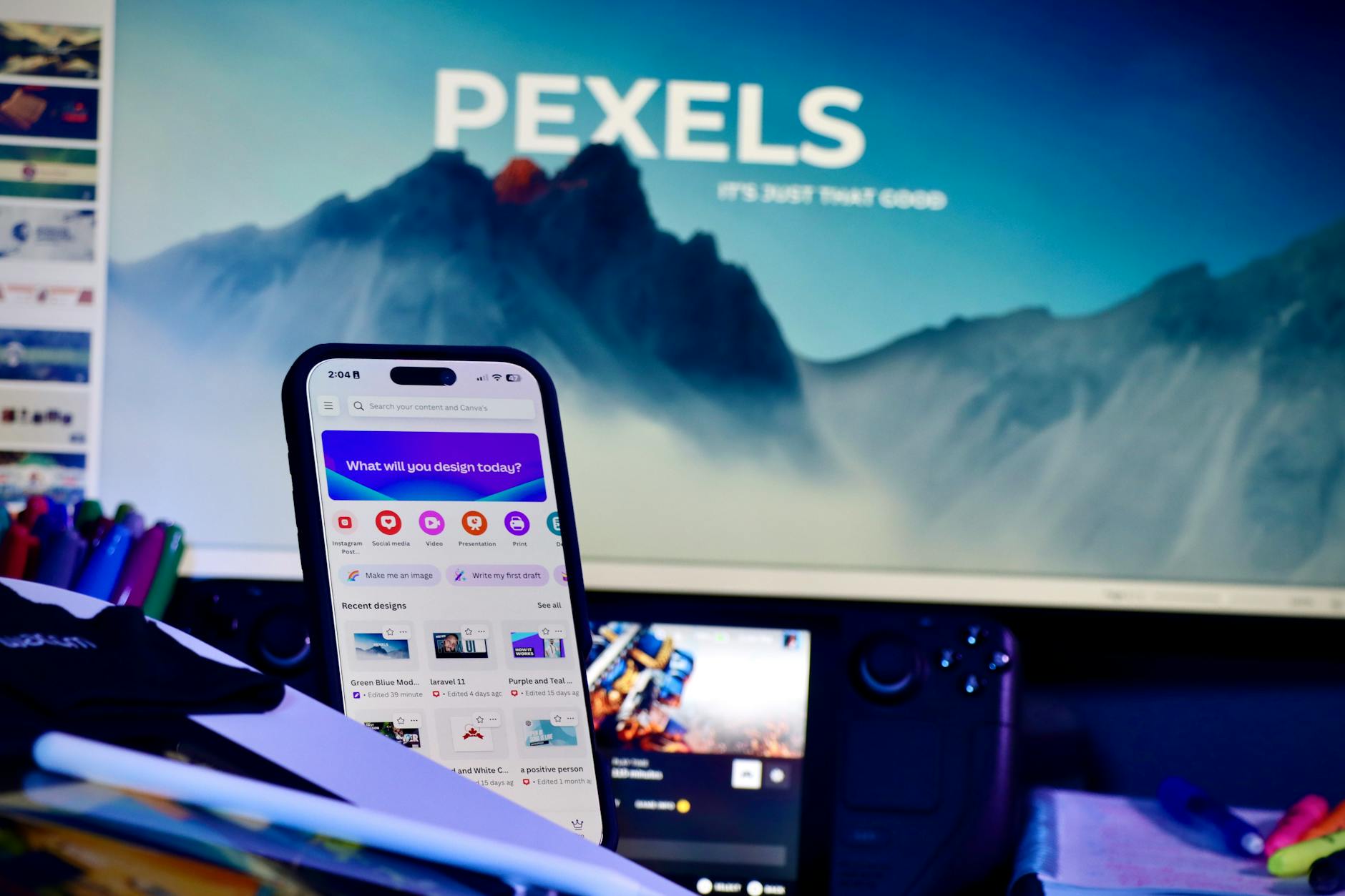 A creative workspace with a smartphone displaying design apps and a large monitor in the background.