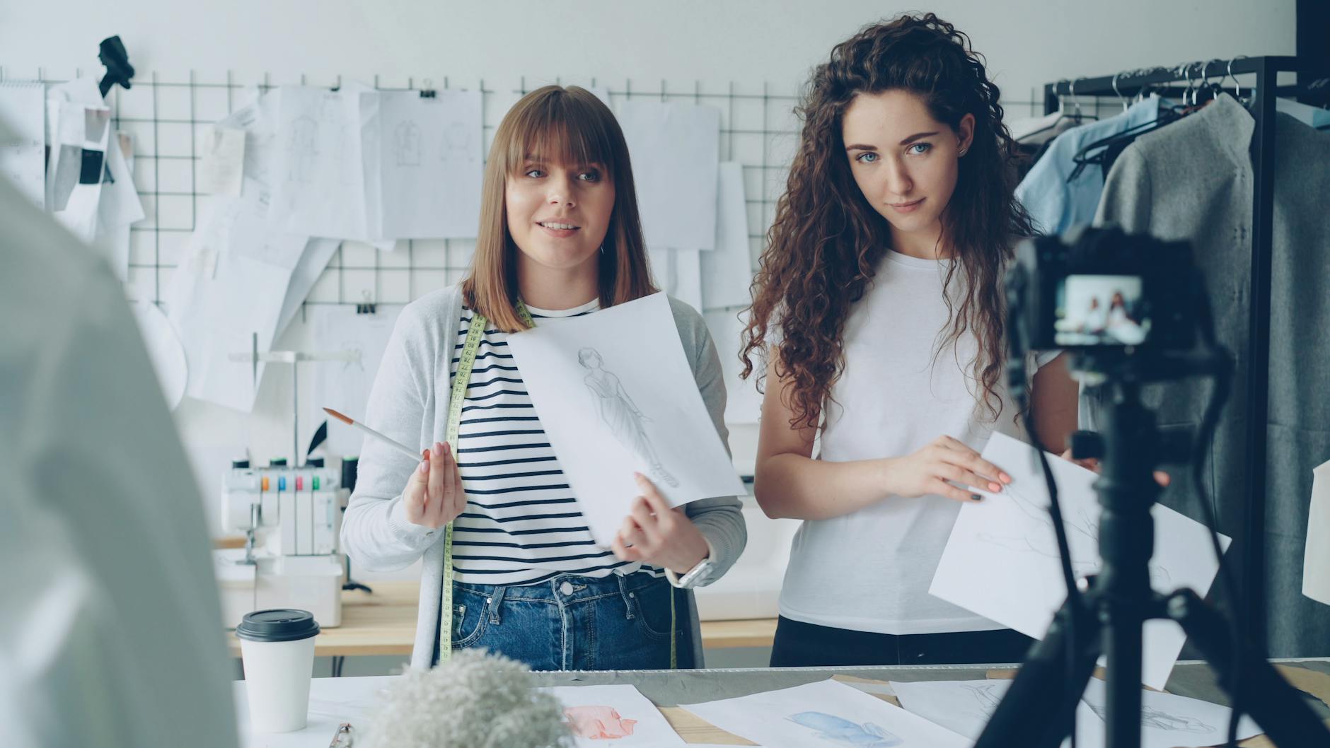 Two female fashion designers showcasing sketches in a design studio with recorded video.