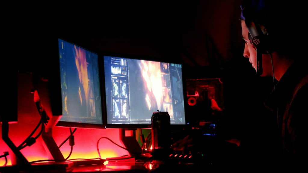 A man intensely focused on multiple computer monitors in a dimly lit room, illustrating high-tech work environment.