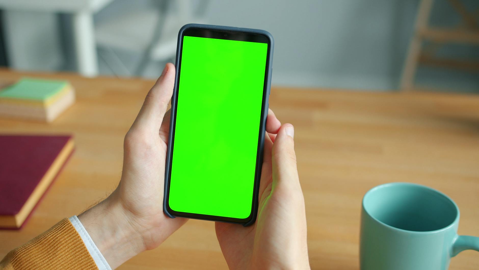Close-up of a person holding a smartphone with a green screen and a mug on a wooden table.
