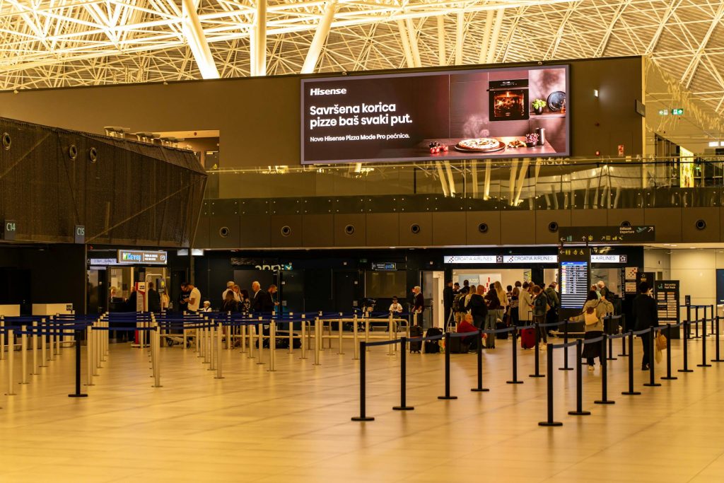 Travelers check in at Zagreb Airport with large advertising screen above.