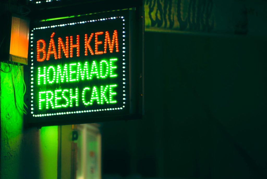 Vibrant neon sign for Bánh Kem displaying "Homemade Fresh Cake" in a dark background.
