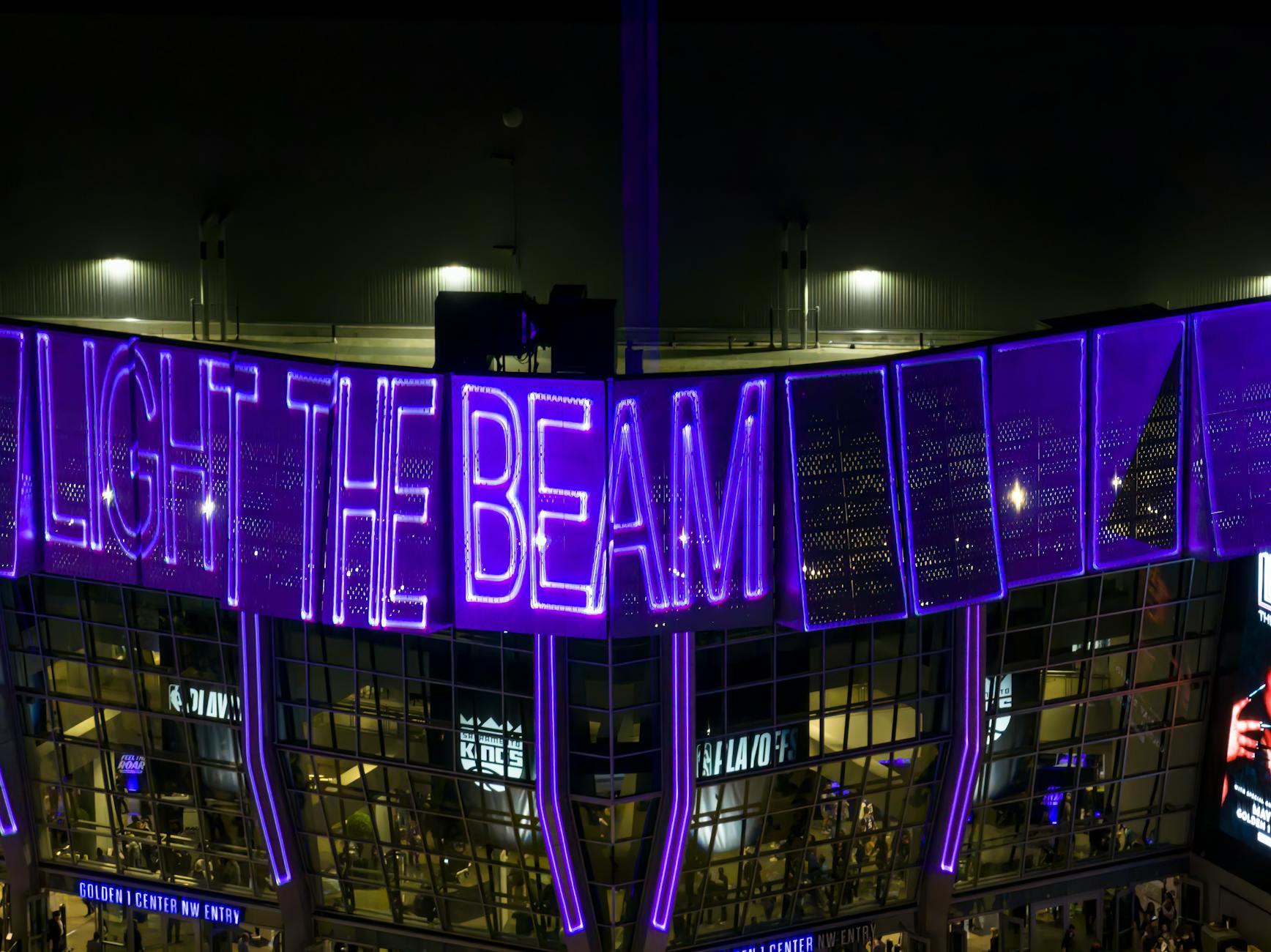 Nighttime view of Golden 1 Center with glowing 'Light the Beam' signage, capturing its vibrant atmosphere.