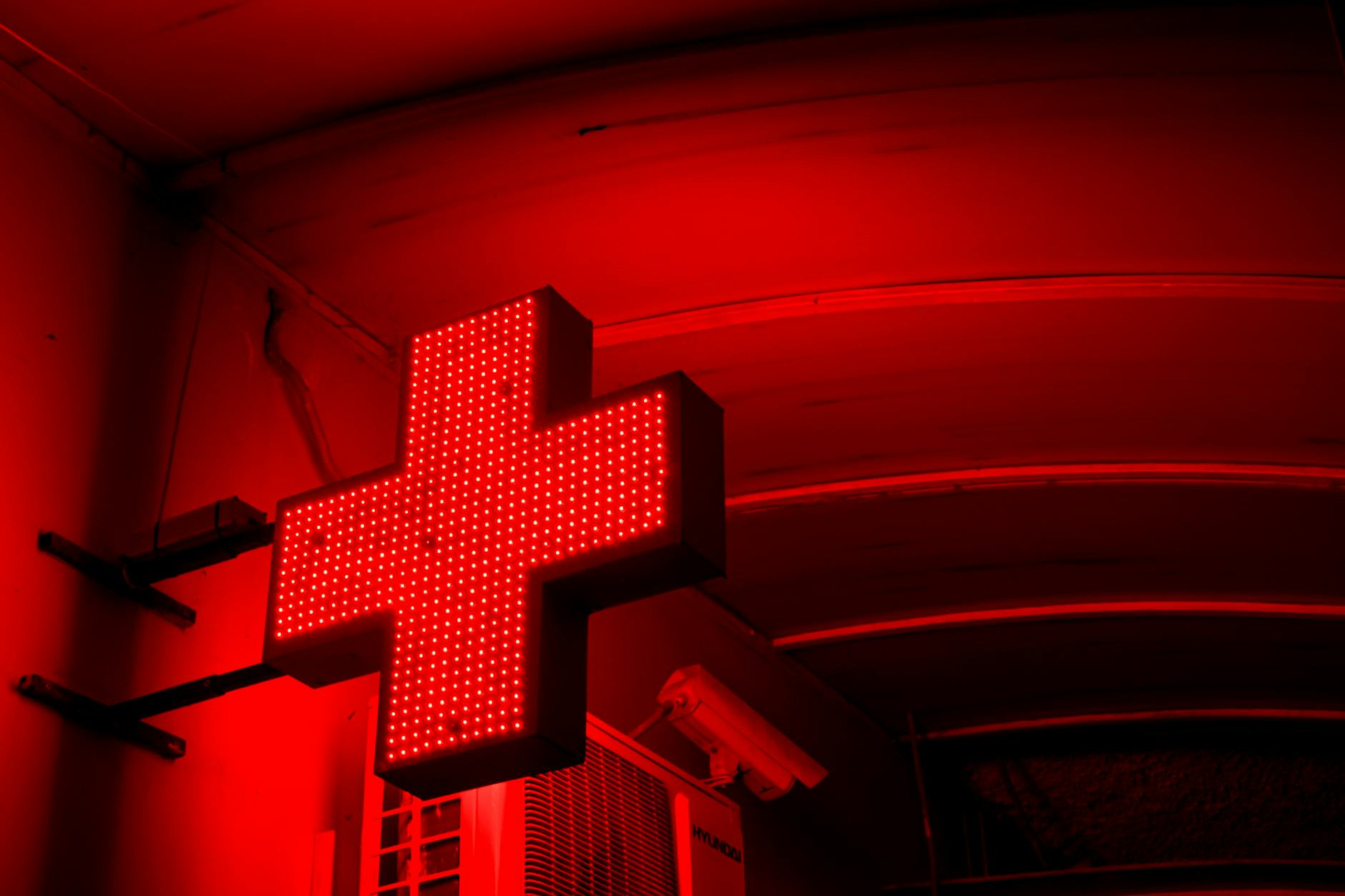 A vibrant red cross sign glowing brightly in a nighttime urban setting.