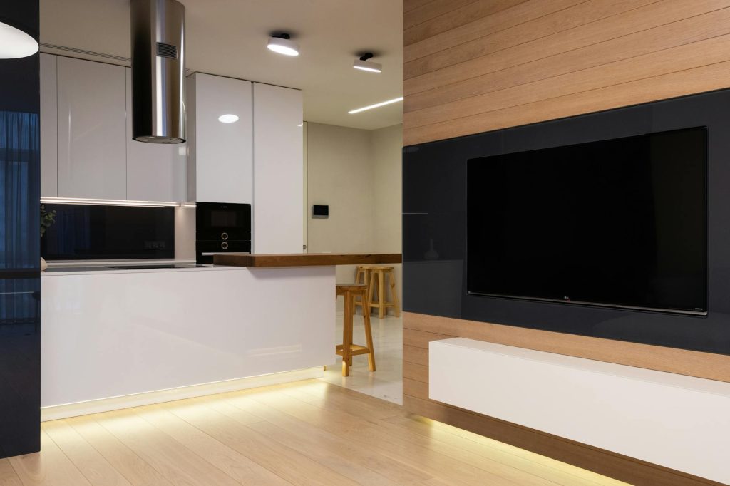 Modern flat with TV set hanging on wall in living room and kitchen furnished with glossy cabinets and shining lamps