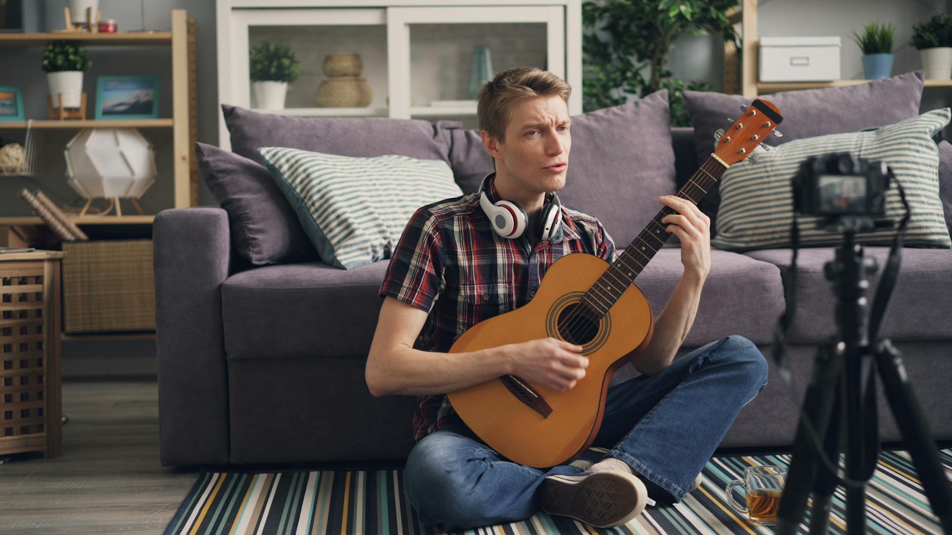 Man sitting cross-legged playing acoustic guitar indoors, streaming online.