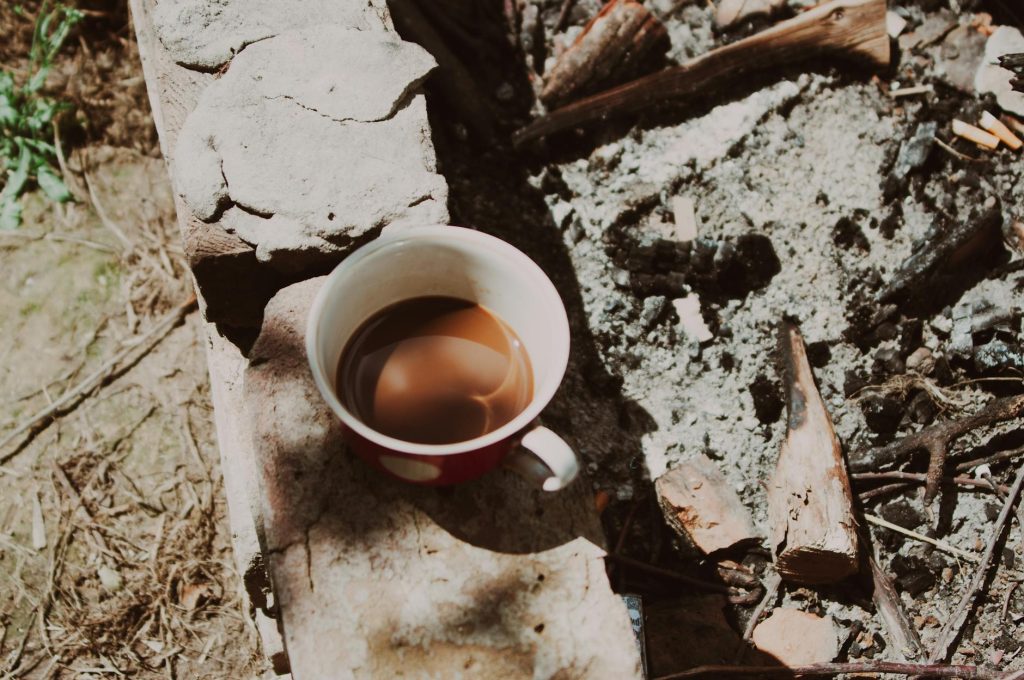 A warm coffee cup sits by a rustic fire pit surrounded by nature, invoking a cozy outdoor vibe.