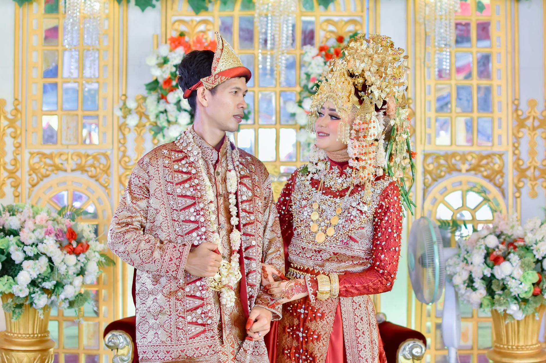 Smiling Indonesian bride and groom in intricate traditional wedding attire, adorned with jewelry and embroidery.