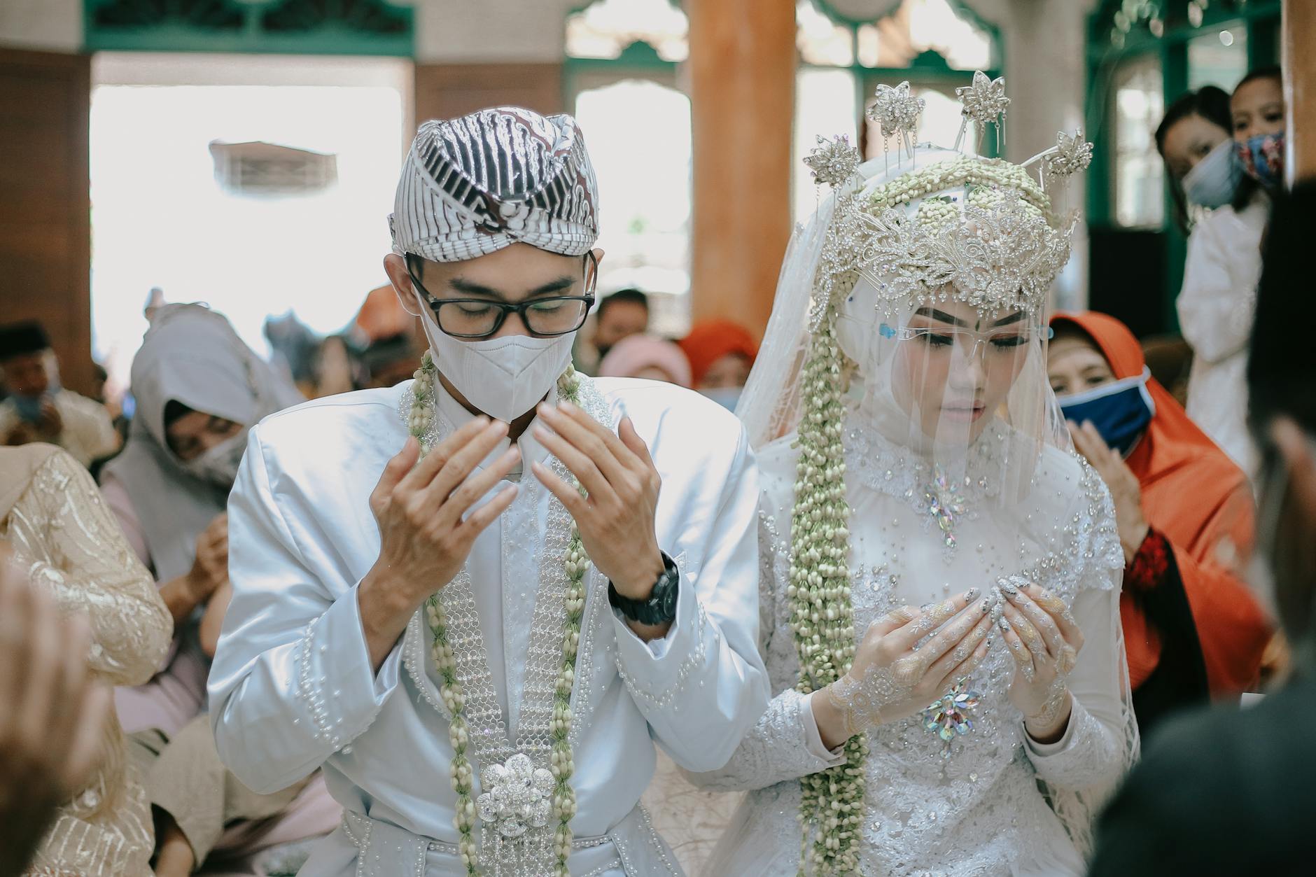 A couple in traditional attire during a wedding ceremony, praying with guests.