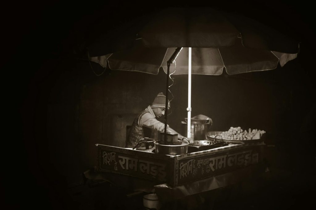A grayscale photo of an Indian street vendor selling food at night under an umbrella.