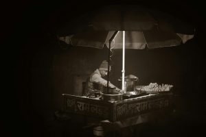 A grayscale photo of an Indian street vendor selling food at night under an umbrella.