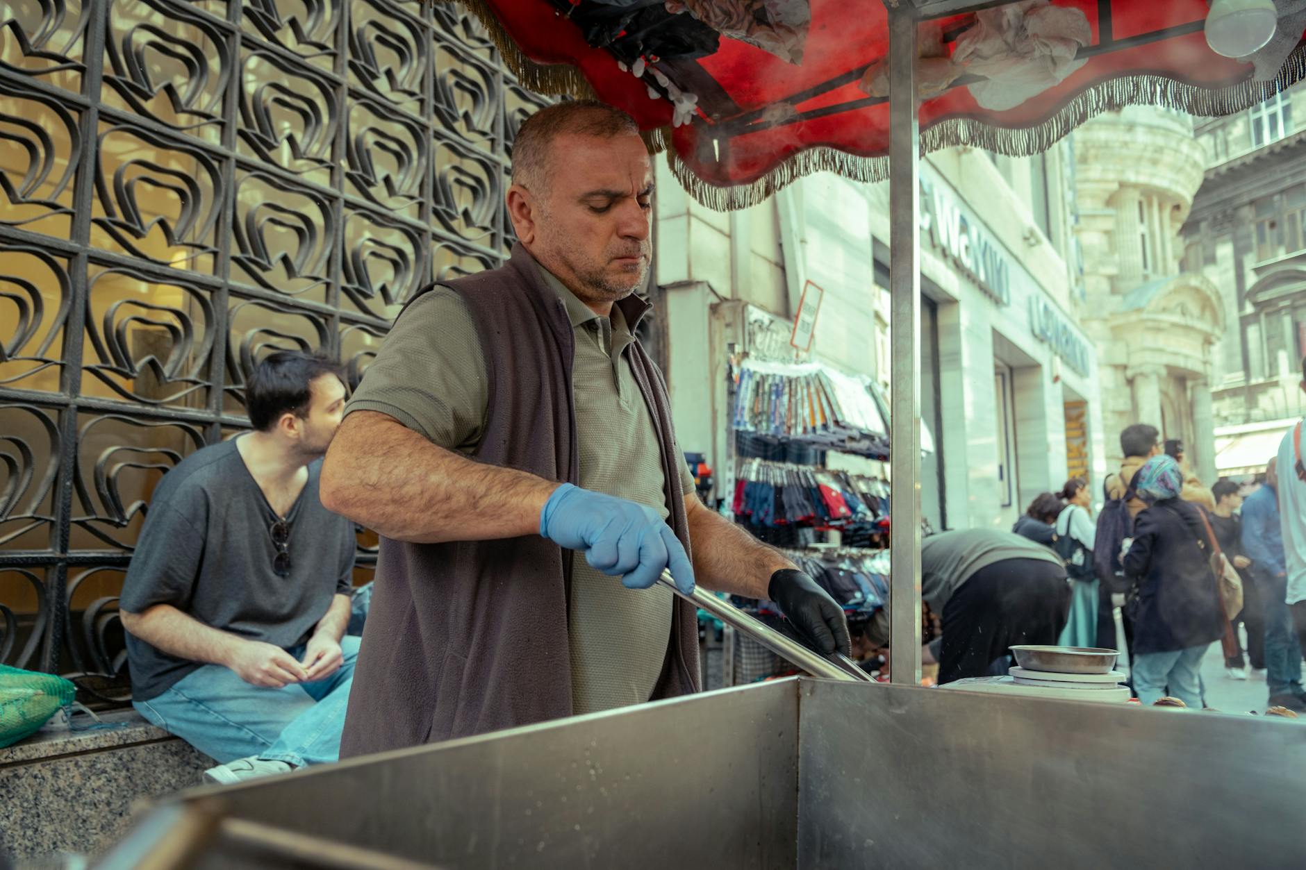 A street vendor in gloves prepares food at an urban market with shoppers nearby, showcasing city life.