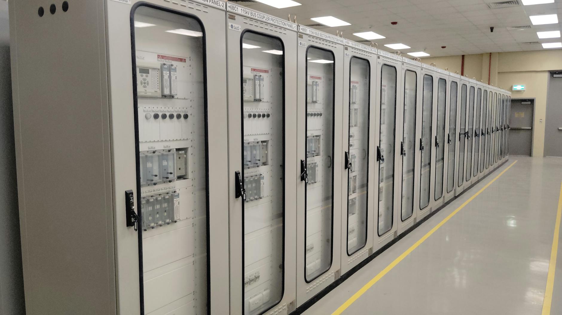 View of an industrial control room featuring rows of electrical panels in a well-lit environment.