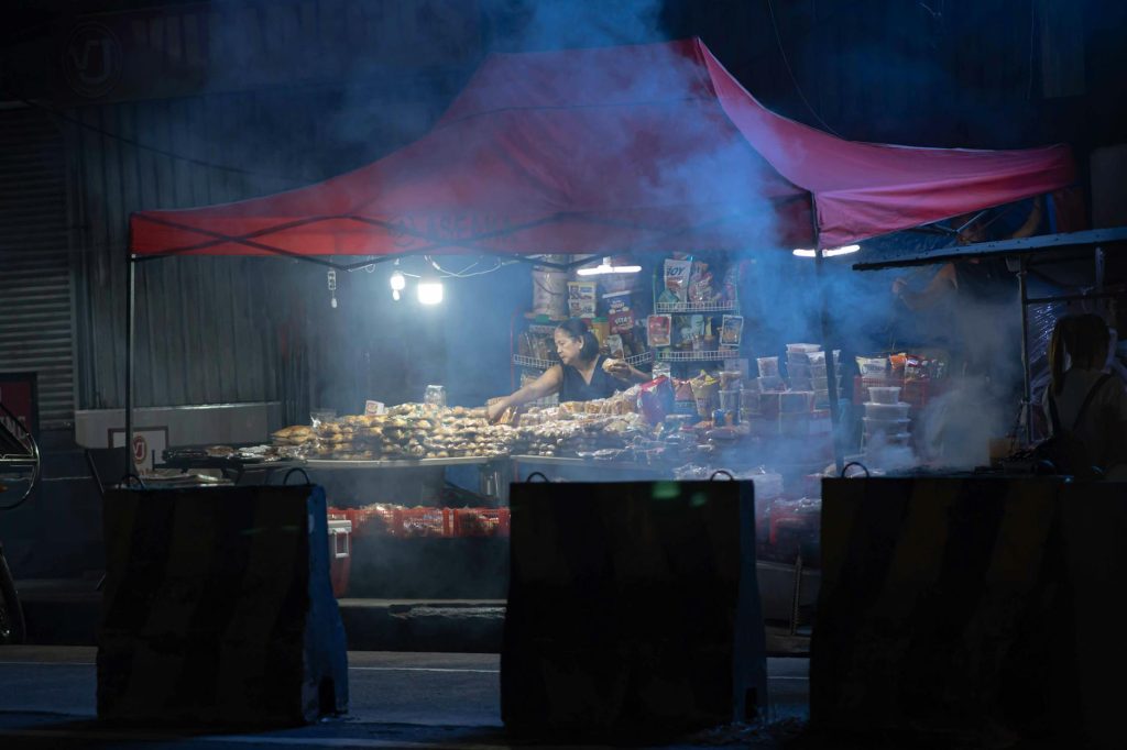 A vendor under a lit canopy selling street food at night with smoke creating a mysterious ambiance.