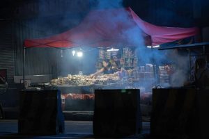 A vendor under a lit canopy selling street food at night with smoke creating a mysterious ambiance.