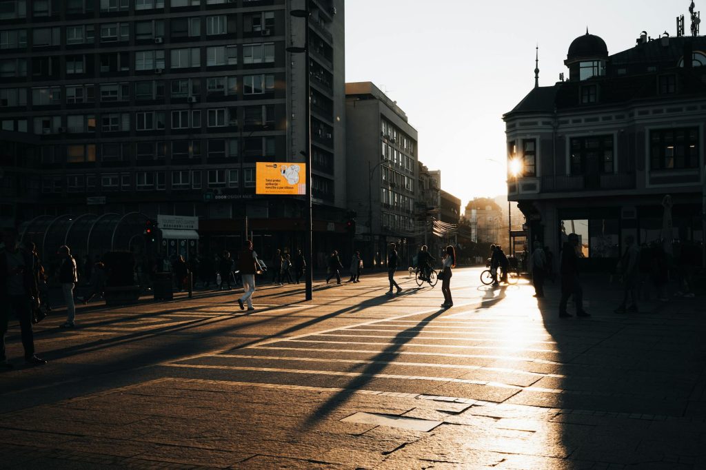 People crossing a city street at sunset, casting long shadows in the warm light.