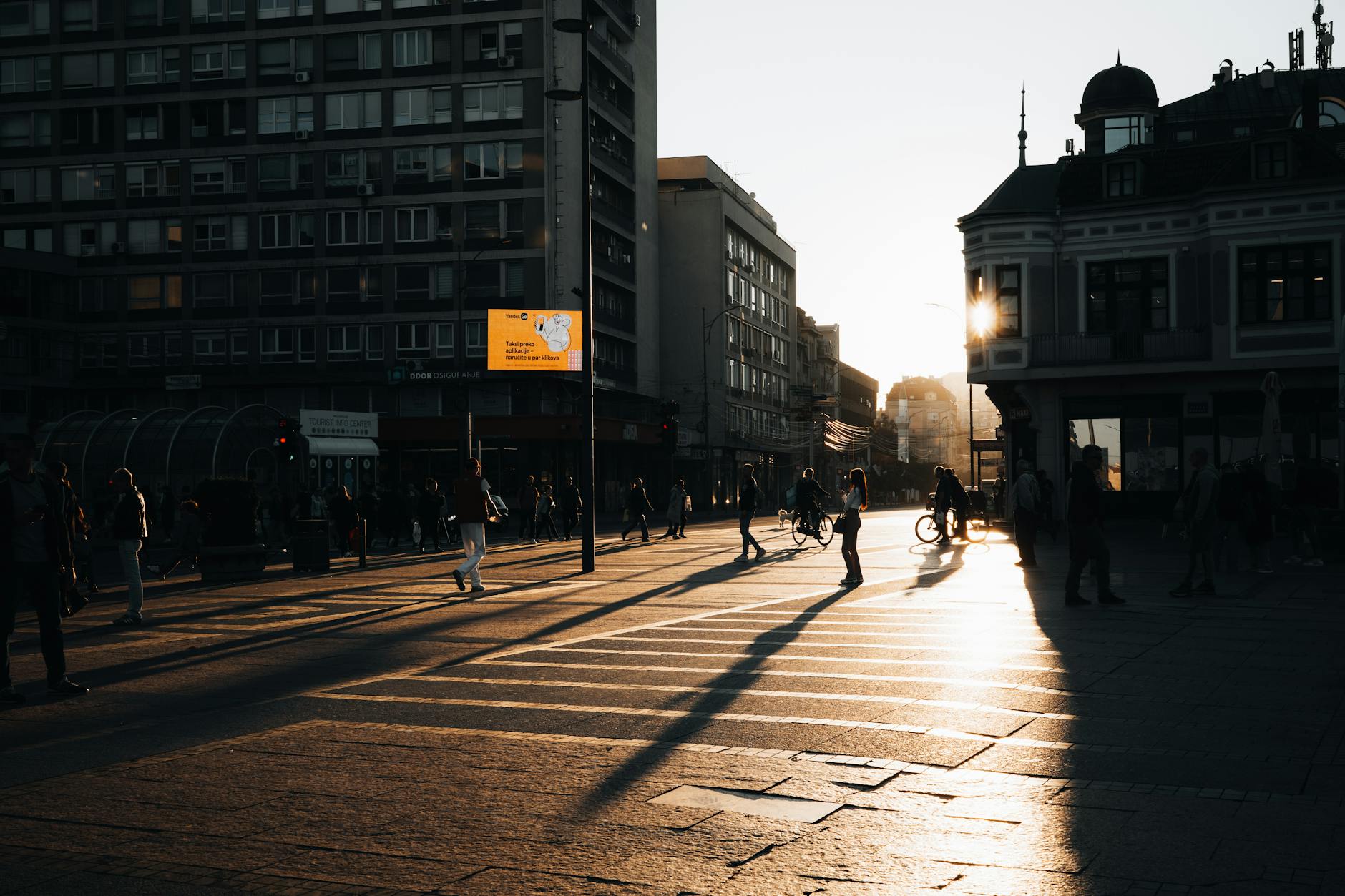 People crossing a city street at sunset, casting long shadows in the warm light.
