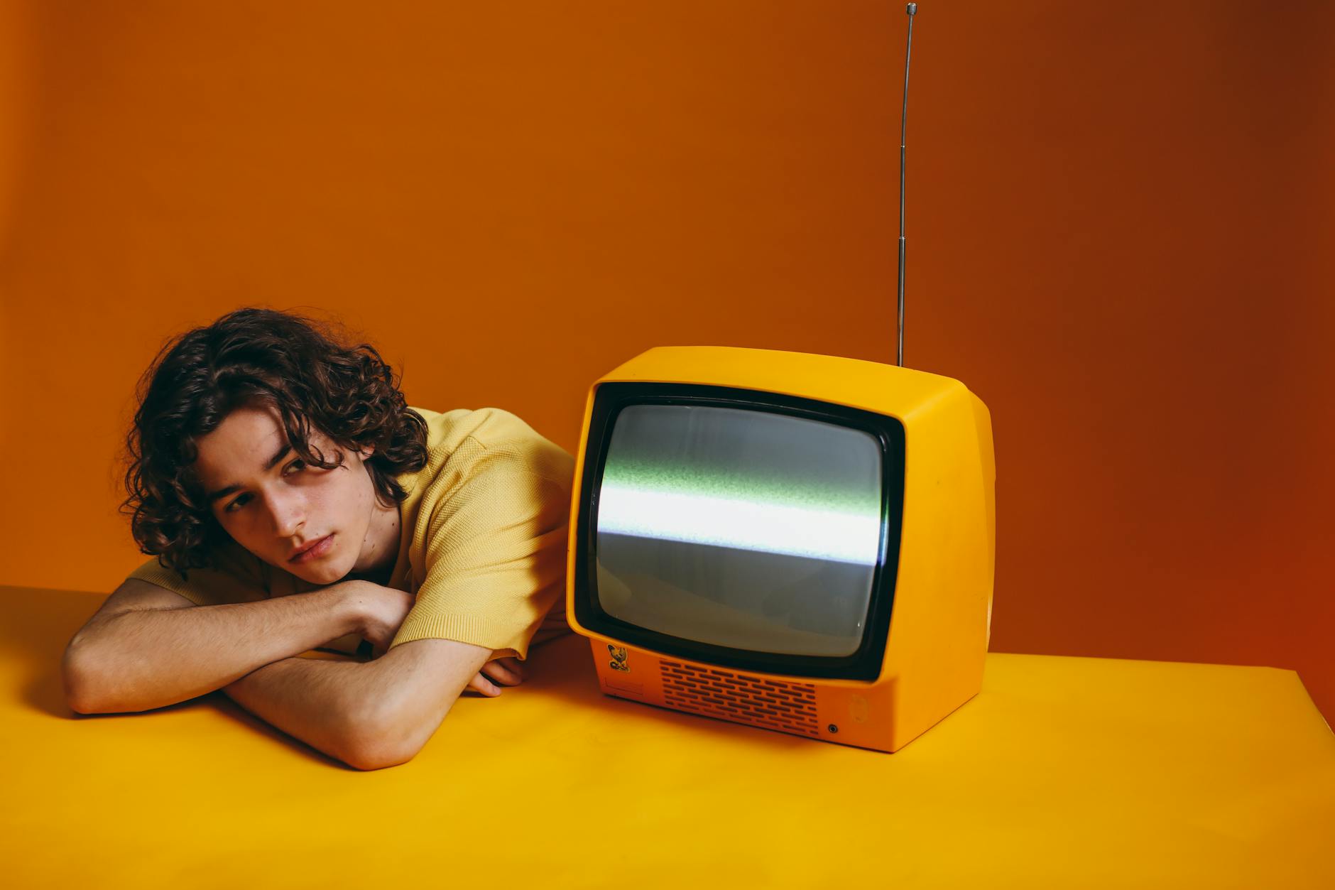 A young man with curly hair rests beside a vintage orange TV on a vibrant backdrop.