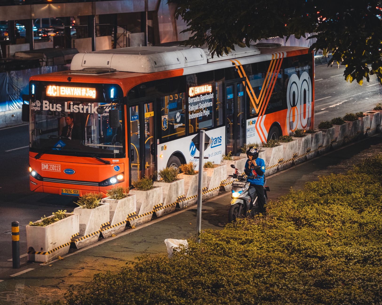 Electric bus and motorcyclist in vibrant Jakarta city at night.