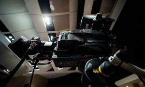 Contemporary professional video camera with lens placed on tripod under ceiling with lights in modern studio