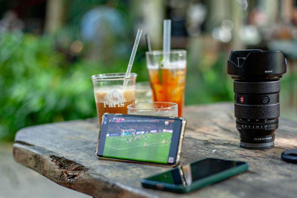 A smartphone streams soccer beside drinks and camera gear at an outdoor cafe.