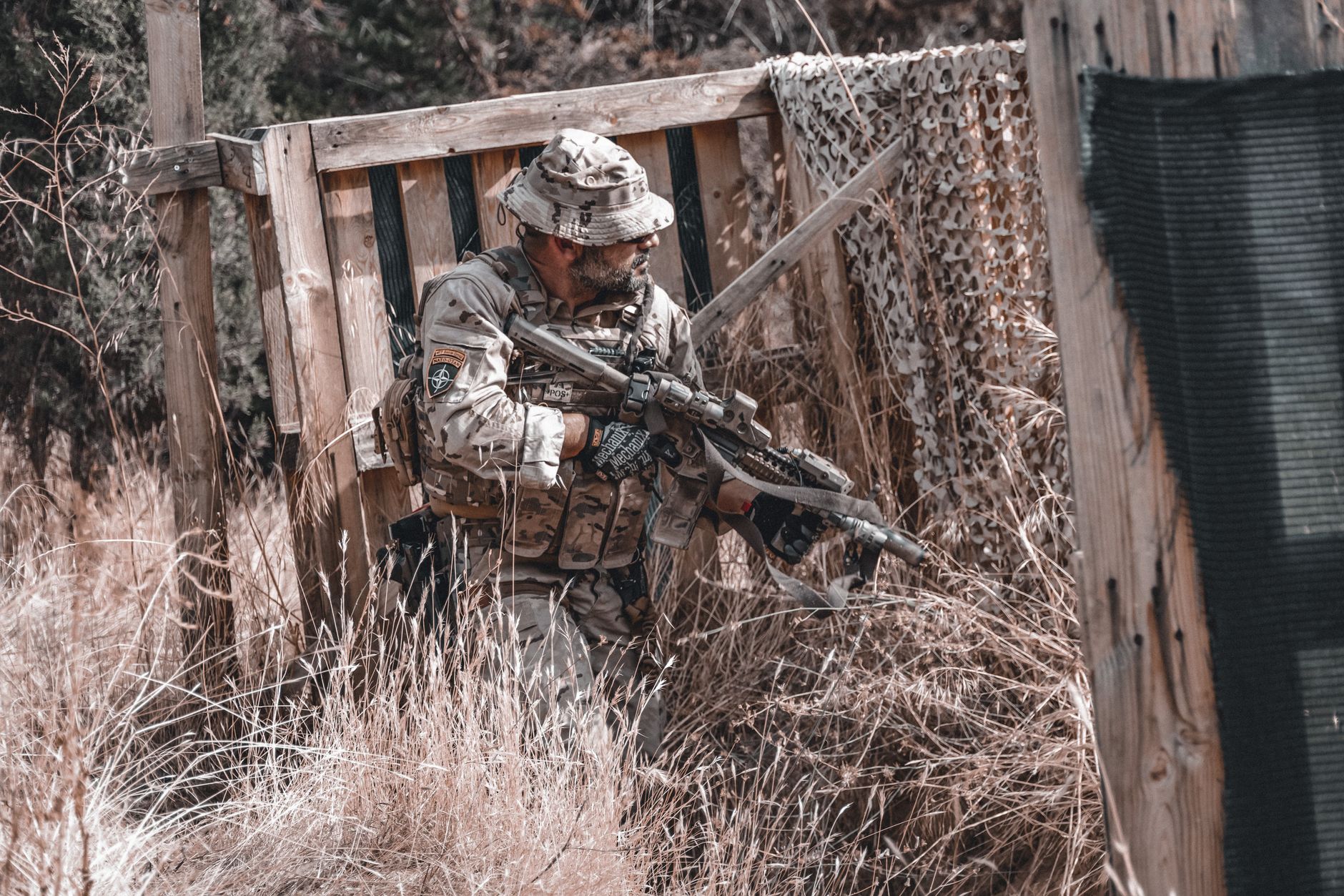 A soldier in full camouflage gear holding a rifle, hiding in an outdoor setting, Spain.