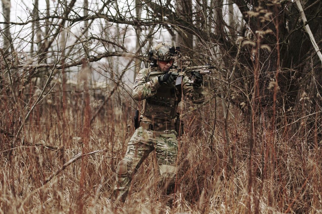 Armed soldier in camouflage attire moving through a dense forest during autumn.