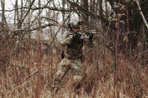 Armed soldier in camouflage attire moving through a dense forest during autumn.