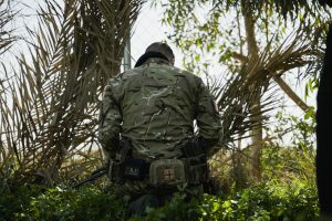 A soldier in camouflage kneels outdoors, focused on surveillance near a fence surrounded by foliage.