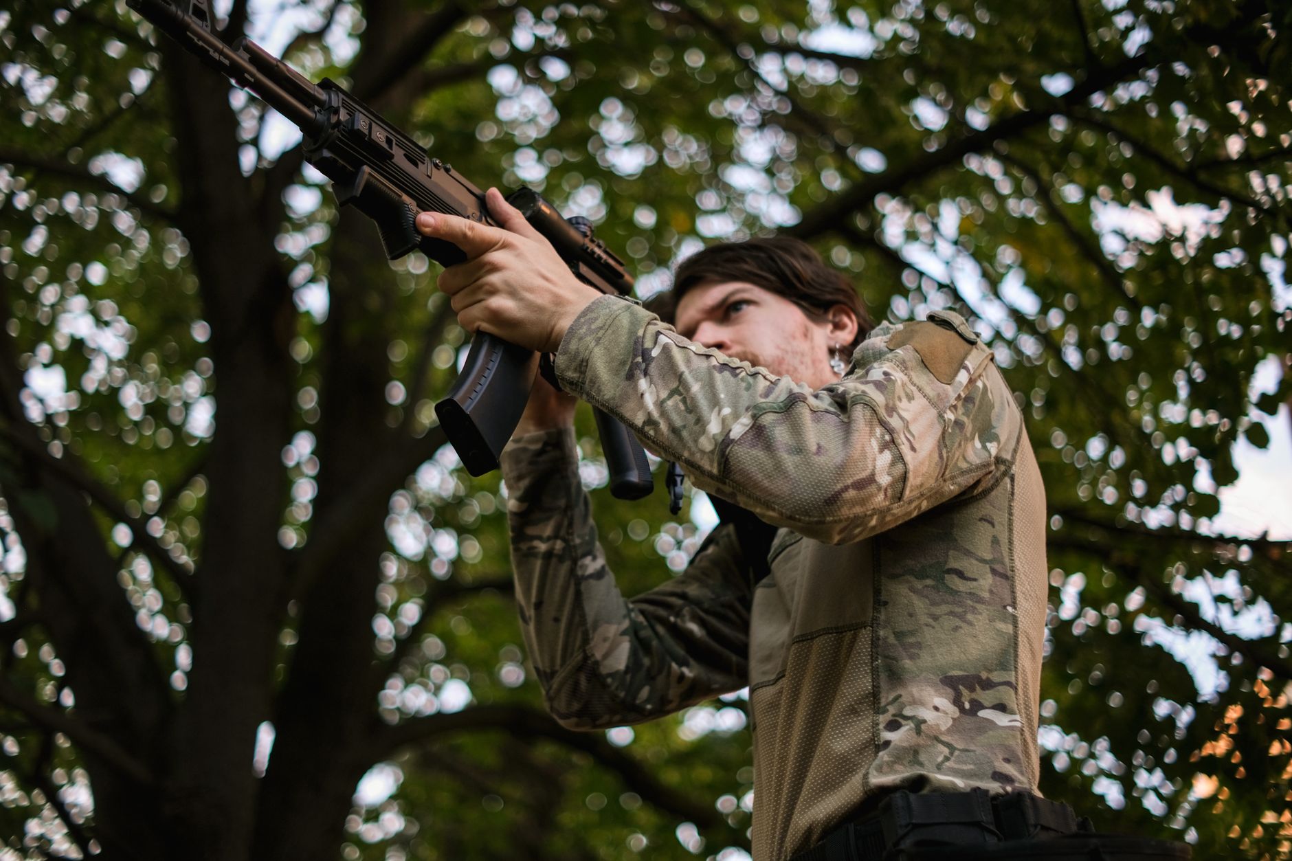 Person in camo gear aims airsoft rifle in wooded area, ready for action.