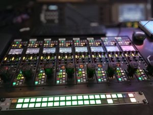 Close-up of an illuminated audio mixer panel in a recording studio, showcasing various controls and switches.
