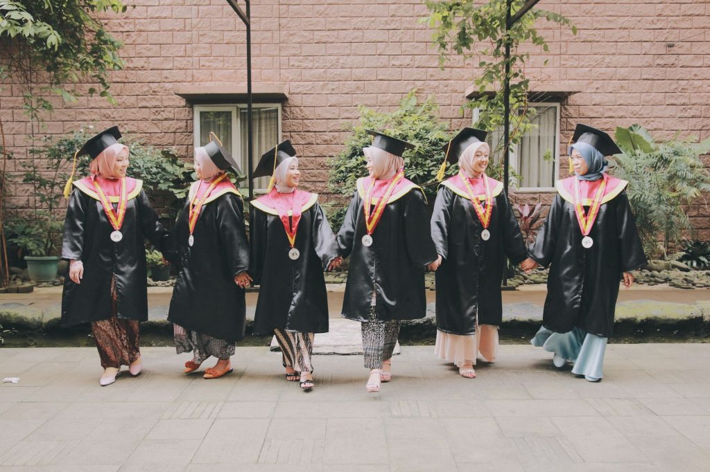 Six female graduates in hijabs and academic gowns celebrating outdoors, smiling and holding hands.