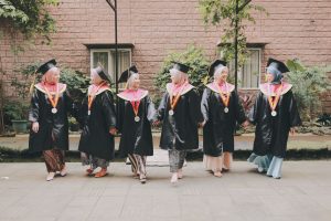 Six female graduates in hijabs and academic gowns celebrating outdoors, smiling and holding hands.