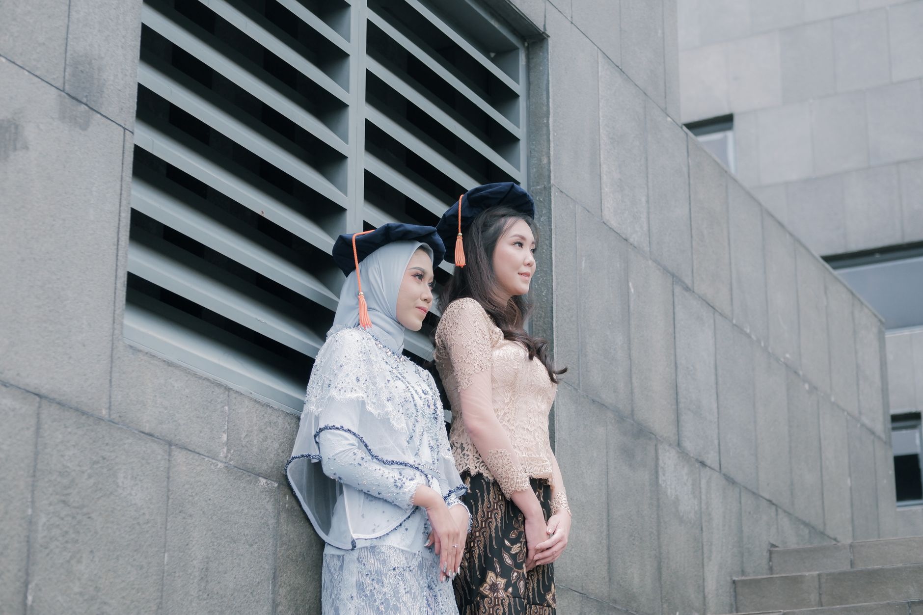 Two women in urban setting wearing traditional attire and graduation caps, reflecting academic achievement.
